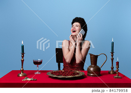 Portrait of young adorable gorgeous woman in dark red, wine color evening dress talking on phone isolated on blue background. 84179958