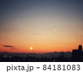夕暮れの太陽と三日月が浮かぶオレンジの空と街並みの風景 84181083