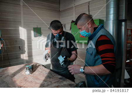 Industrial work during a pandemic. Two men work in a heavy metal factory, wearing a mask on their face due to a coronavirus pandemic Industrial work during a pandemic. Two men work in a heavy metal factory, wearing a mask on their face due to a coronavirus pandemic 84191817