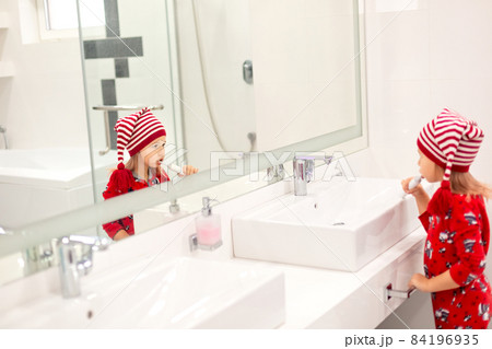 little girl in santa claus hat and red pajama brushing teeth in white bathroom in the morning on Christmas holidays 84196935