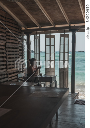 Girl sitting and drinking coffee in a wooden cafe overlooking the ocean. Blue crystal clear water in the ocean. Lovely view from the coffee shop Girl sitting and drinking coffee in a wooden cafe overlooking the ocean. Blue crystal clear water in the ocean. Lovely view from the coffee shop 84200950