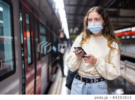 Young woman in mask using mobile phone while waiting metro 84201613