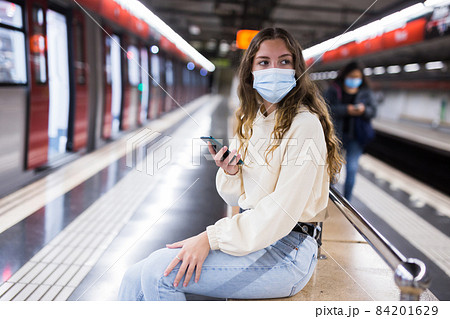 Girl in a protective mask sitting on a bench in the subway with a mobile phone waiting for a train 84201629