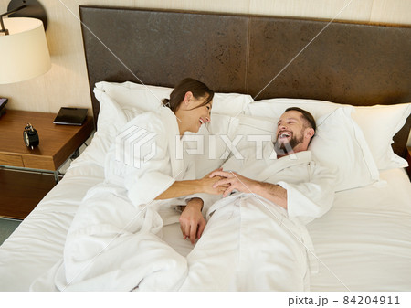 Charming couple in love of a beautiful woman and a handsome man, happy husband and wife, newlyweds, dressed in terry bathrobe, spending time relaxing in bed, enjoying the privacy of their honeymoon. Charming couple in love of a beautiful woman and a handsome man, happy husband and wife, newlyweds, dressed in terry bathrobe, spending time relaxing in bed, enjoying the privacy of their honeymoon. 84204911