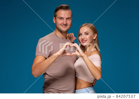 Happy couple dressed casual clothes, making heart shape from fingers, studio portrait on blue background Happy couple dressed casual clothes, making heart shape from fingers, studio portrait on blue background 84207537