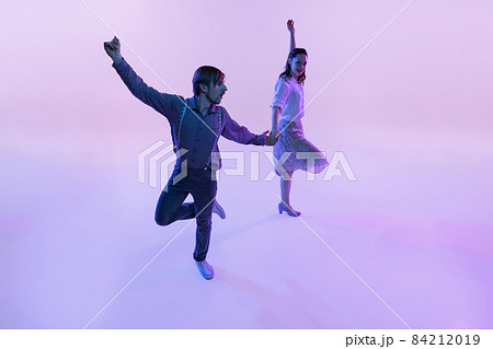 Studio shot of young man and woman in vintage retro style outfits dancing lindy hop isolated on lilac color background in neon light Studio shot of young man and woman in vintage retro style outfits dancing lindy hop isolated on lilac color background in neon light 84212019