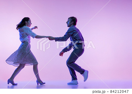 Two young graceful dancers, man and woman in vintage style outfits dancing rock-and-roll isolated on lilac color background in neon light 84212047