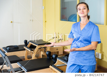 Smiling female physiotherapist inviting to rehab center with pilates equipment Smiling female physiotherapist inviting to rehab center with pilates equipment 84214747