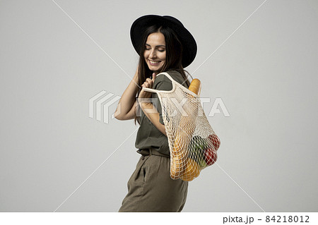 Zero waste concept. A young girl holds on her shoulder a textile mesh eco bag with a groceries. The girl smiles, wearing a beige t-shirt and hat. Refusal of plastic bags. 84218012
