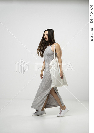 Brunette woman in grey dress holding cotton shopper bag with vegetables, products in white room. Eco friendly shopping bags. Zero waste, plastic free concept. 84218074