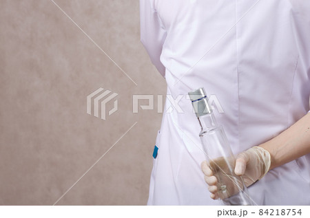 A young woman dressed in a medical uniform hides a bottle of alcohol. A young woman dressed in a medical uniform hides a bottle of alcohol. 84218754