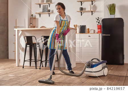 Young woman using vacuum cleaner in home kitchen floor, doing cleaning duties and chores, meticulous interior. 84219657