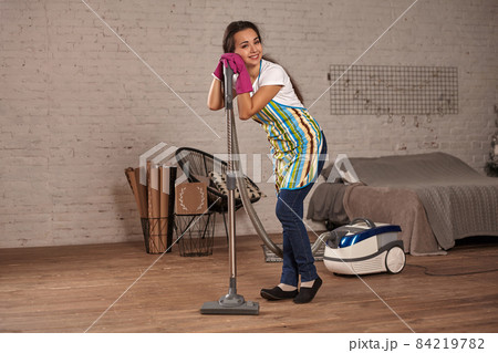 Young woman using vacuum cleaner in home living room floor, doing cleaning duties and chores, meticulous interior. 84219782