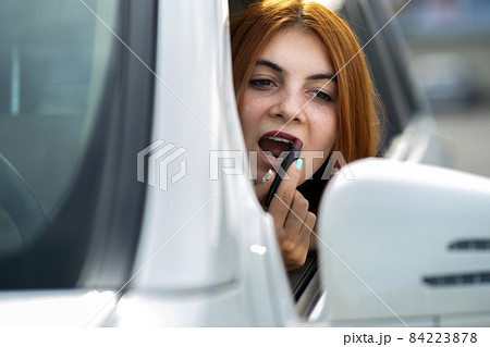 Closeup of a young redhead woman driver correcting her makeup with dark red lipstick looking in car rearview mirror behind steering wheel of a vehicle. Closeup of a young redhead woman driver correcting her makeup with dark red lipstick looking in car rearview mirror behind steering wheel of a vehicle. 84223878