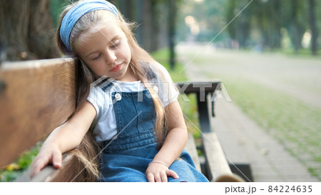 Small tired child girl sitting on a bench with closed eyes resting in summer park. Small tired child girl sitting on a bench with closed eyes resting in summer park. 84224635