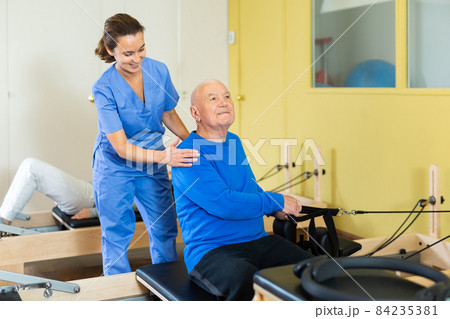 Medical professional helps and monitors the difficulty of Pilates exercise for an mature man Medical professional helps and monitors the difficulty of Pilates exercise for an mature man 84235381