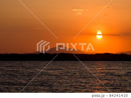 涸沼の日没。オレンジ色の空に太陽が輝き、その太陽を一本の矢が射貫く様に雲が伸びる。その下に筑波山 84241410