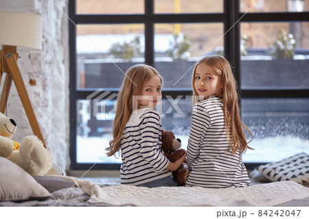 The image of two little sisters sitting on the bed in the room. 84242047