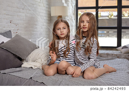 The image of two little sisters sitting on the bed in the room. The image of two little sisters sitting on the bed in the room. 84242493