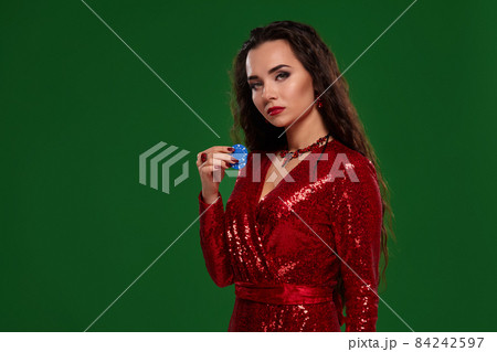 Young pretty lucky brunette woman in a red evening dress holds few poker chips in her hands on green background 84242597