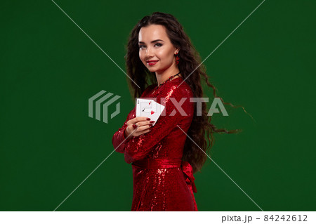 Young beautiful lucky brunette in a red evening dress shows her cards, a pair of aces on green background Young beautiful lucky brunette in a red evening dress shows her cards, a pair of aces on green background 84242612