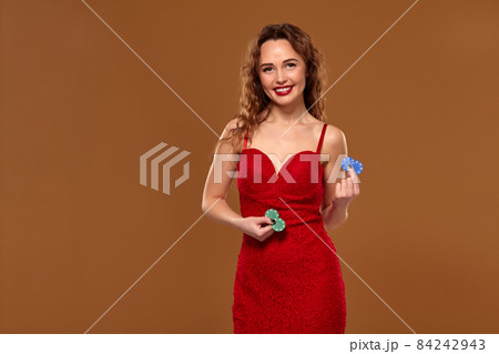 Blond curly woman weared in a red sexy dress is holding some blue gambling chips in her hands. Casino. Blond curly woman weared in a red sexy dress is holding some blue gambling chips in her hands. Casino. 84242943