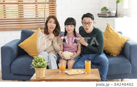 Excited Asian family eating popcorn and watching tv together on sofa at home living room. leisure and people concept.. Excited Asian family eating popcorn and watching tv together on sofa at home living room. leisure and people concept.. 84248402