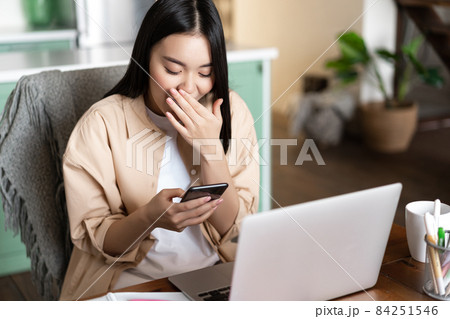 Happy asian girl sitting with laptop at home, looking at mobile phone and laughing, has workplace at house 84251546