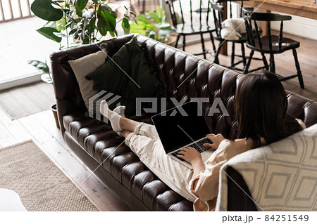 Rear view of young woman relaxing at home with laptop. Girl sits on couch with computer, watching videos or browsing websites Rear view of young woman relaxing at home with laptop. Girl sits on couch with computer, watching videos or browsing websites 84251549