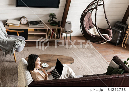 Portrait of young woman sitting at home with laptop. Asian girl using computer pc in her living room, resting on sofa Portrait of young woman sitting at home with laptop. Asian girl using computer pc in her living room, resting on sofa 84251738