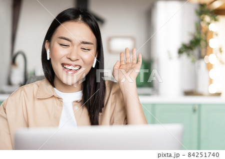 Happy asian woman laughing and smiling, talking on video call using laptop, waving hand and chuckling, wearing earbuds Happy asian woman laughing and smiling, talking on video call using laptop, waving hand and chuckling, wearing earbuds 84251740
