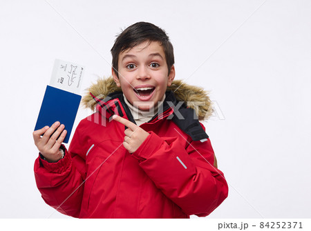 Beautiful preadolescent boy, cheerful school child in bright red warm parka points with his finger at a passport with a ticket and boarding pass, isolated over white background with copy ad space 84252371