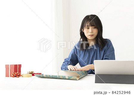 窓の近くのテーブルで包み方をネットで検索しながらプレゼントを包装紙で包む女性 84252946