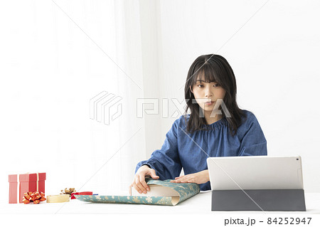 窓の近くのテーブルで包み方をネットで検索しながらプレゼントを包装紙で包む女性 84252947