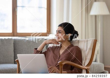 Woman relaxing on armchair with laptop staring into distance daydreaming Woman relaxing on armchair with laptop staring into distance daydreaming 84259397