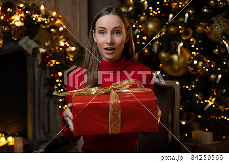 Happy woman in a red sweater opens a magical Christmas gift at home on the background of a Christmas tree 84259566