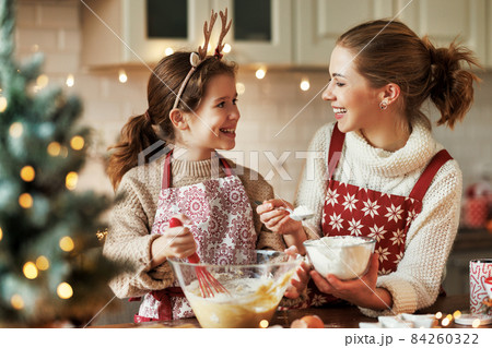 Cute little daughter girl helping mother to make dough for Christmas cookies in cozy kitchen at home Cute little daughter girl helping mother to make dough for Christmas cookies in cozy kitchen at home 84260322