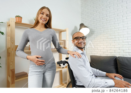 Pregnant woman holding a hand of her disabled husband sitting in a wheelchair 84264186