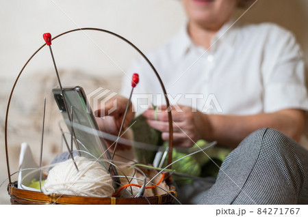 Faceless old woman knits and watches online training on a smartphone. Close-up of female hands with yarn and knitting needles. Faceless old woman knits and watches online training on a smartphone. Close-up of female hands with yarn and knitting needles. 84271767