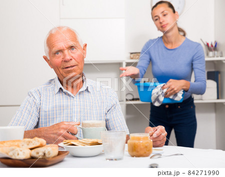 Mature father drinking tea in the kitchen. Adult daughter does cleaning in the apartment Mature father drinking tea in the kitchen. Adult daughter does cleaning in the apartment 84271990