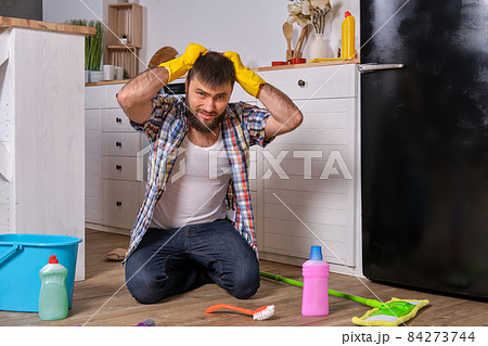 Caucasian young bearded man sits on the floor of his kitchen and tries to clean it using all of his detergents, rags, washcloths and brushes 84273744