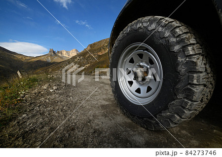 Close-up A large off-road all-terrain car wheel stands on a rough road in the mountains against the backdrop of rocks. Off-road tours and off-road travel concept 84273746