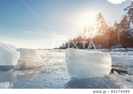 Scenic view of clear ice shard piece on coast of frozen lake landscape against warm sun shining rays through forest tree and clear blue sky on background. Winter weather nature abstract scene view Scenic view of clear ice shard piece on coast of frozen lake landscape against warm sun shining rays through forest tree and clear blue sky on background. Winter weather nature abstract scene view 84274099