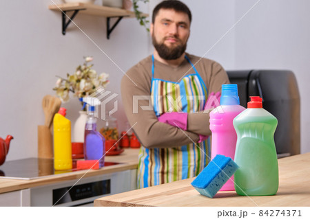 Presentation of different detergents with washcloth on the foreground, caucasian bearded man is standing in the background with his arms crossed 84274371