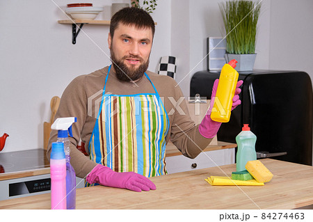 Young handsome bearded man in the kitchen, cleans up using detergents, brushes, sprays 84274463