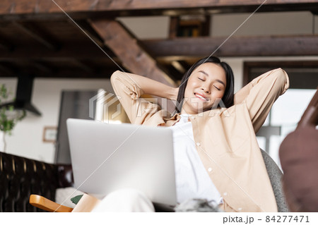 Smiling korean woman resting with hands behind her head, using laptop and relaxing at home 84277471