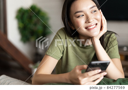 Young asian woman placing an order, waiting for delivery on mobile phone app, sitting at home and looking outside window Young asian woman placing an order, waiting for delivery on mobile phone app, sitting at home and looking outside window 84277569