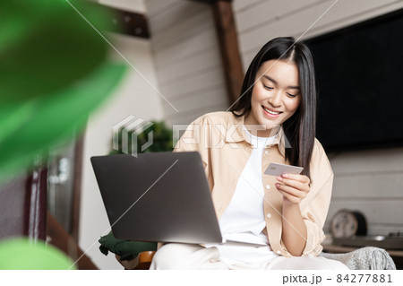 Smiling asian girl paying for online shopping on laptop, looking at her credit card, sitting at home in living room with computer Smiling asian girl paying for online shopping on laptop, looking at her credit card, sitting at home in living room with computer 84277881