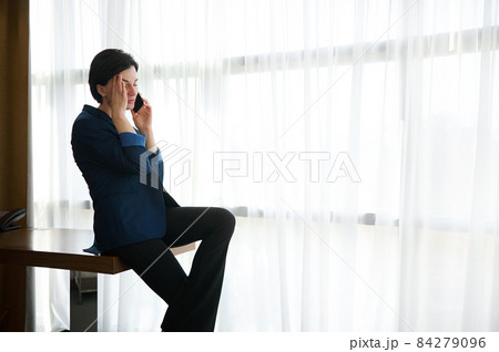 Tired brunette, female entrepreneur, businesswoman at business trip, sitting at the edge of the table by the window in a hotel room and massaging her temple during a boring uninteresting phone call 84279096