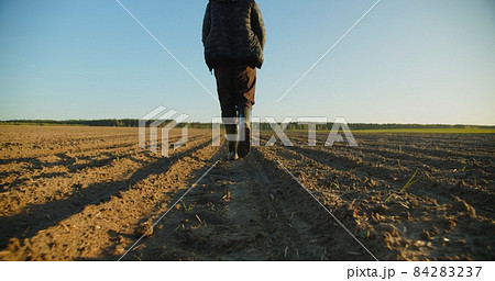Low angle: man walking in rubber boots in a farmer's field, the blue sky above the horizon. Man walking through an agricultural field. Farmer walks through a plowed field in early spring. 84283237